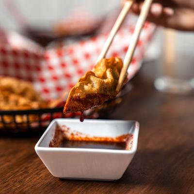 Dumpling held with chopsticks over a small square bowl of dipping sauce.