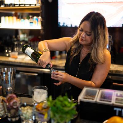 A bartender pouring a drink into a jigger at a bar counter.