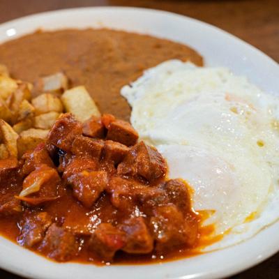 Mexican breakfast plate with eggs, potatoes, refried beans, and meat in red sauce.