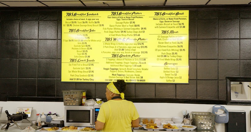 A kitchen staff member standing under a yellow breakfast menu board