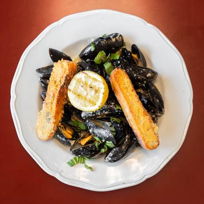 Plate of black mussels topped with garlic breadsticks and lemon slice, garnished with herbs.