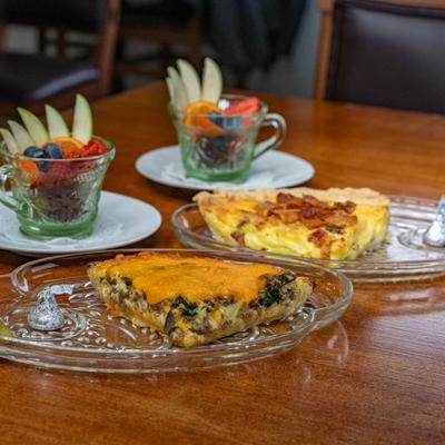 A slice of cheddar and spinach quiche and a slice of bacon quiche served with fruit cups.