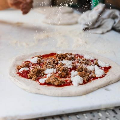 An uncooked meatball pizza being sprinkled with flour.