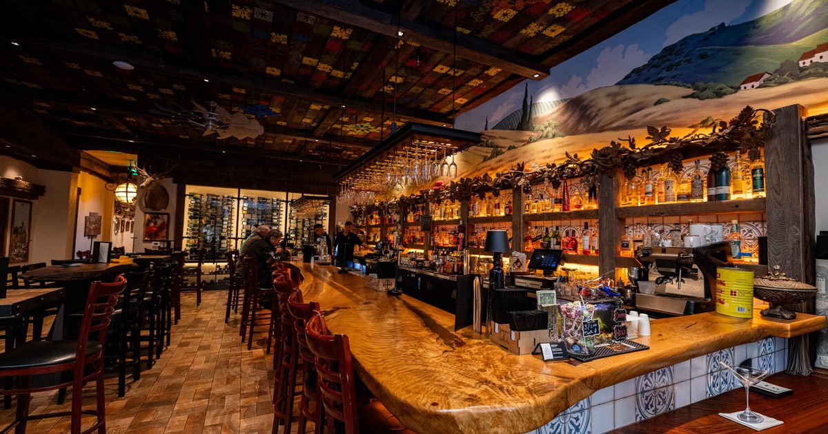 Interior of a warmly lit bar with a long wooden counter, colorful ceiling tiles
