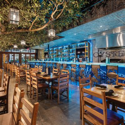 Restaurant interior featuring a blue-lit counter and wooden tables under a large green tree.