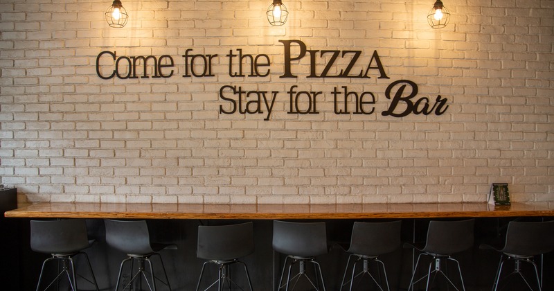 Interior, white brick wall with the writing "Come for the pizza, stay for the bar"