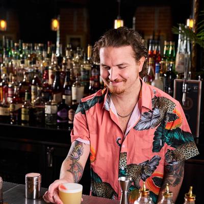 Bartender in a tropical shirt smiles while presenting a foamy yellow cocktail.