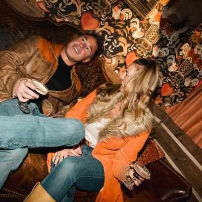 A couple sits closely on a leather couch, holding glasses and smiling.