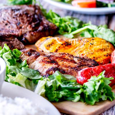 Grilled meats, chicken, and sausages on a wooden board, garnished with fresh greens.