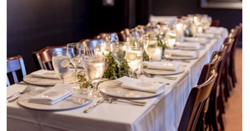 A long table decorated for a dinner event.  White tablecloths, plate-ware, candles and some decorations