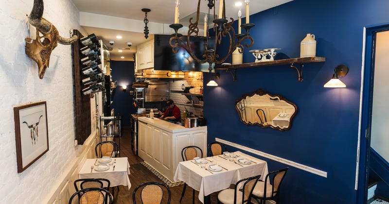 Interior of a small restaurant with white and blue walls, tables set for dining, and a visible kitchen area in the background