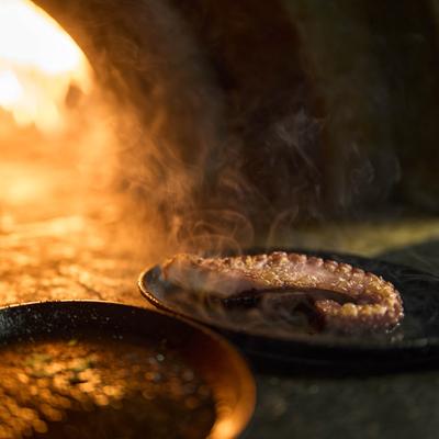 A gourmet dish of grilled octopus being cooked or finished over an open flame.