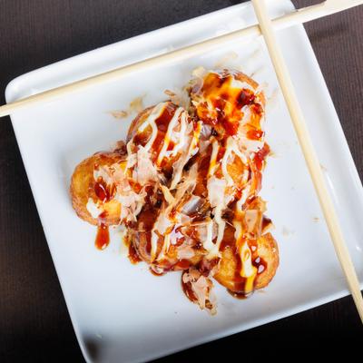 Takoyaki balls drizzled with sauces and bonito flakes on a white plate with chopsticks.