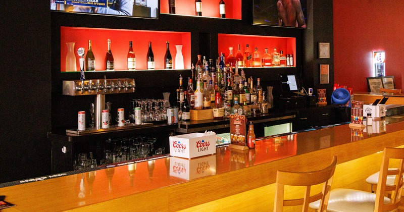 Interior of a bar - a wooden counter, red shelves displaying bottles and two TVs screens