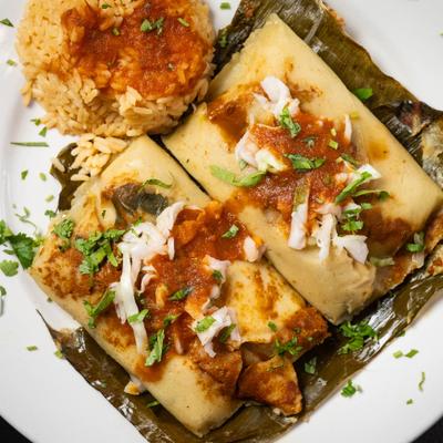 Tamales wrapped in banana leaves served with salsa, cabbage, cilantro, and seasoned rice.