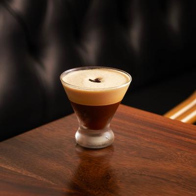 Espresso martini sits on a wooden table with a black booth seating in the background.