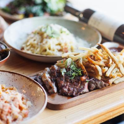 Grilled steak with fries served alongside two plated of pasta and a bottle of wine.