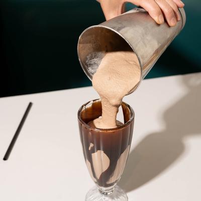 Creamy chocolate milkshake is being poured from a metal shaker into a tall glass.