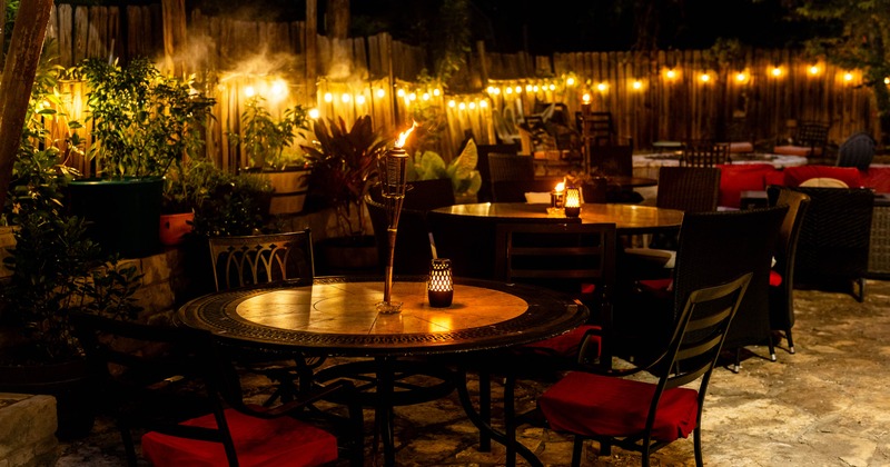 An outdoor dining area in the evening, featuring tables with candles, string lights, and torches