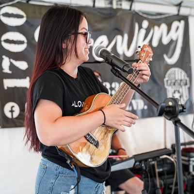 A musician plays ukulele and sings into a microphone at an outdoor music event.