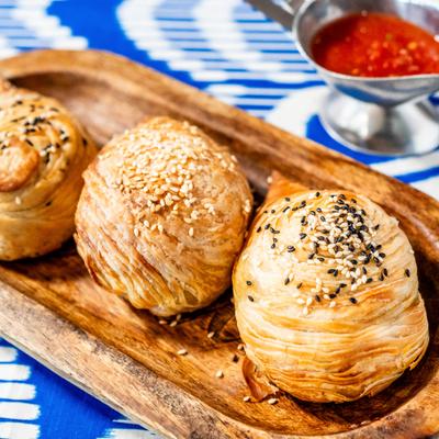 Three flaky sesame pastries on a wooden tray with a side of tomato dipping sauce.