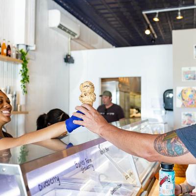 A server hands a smiling customer a double-scoop ice cream across the counter.