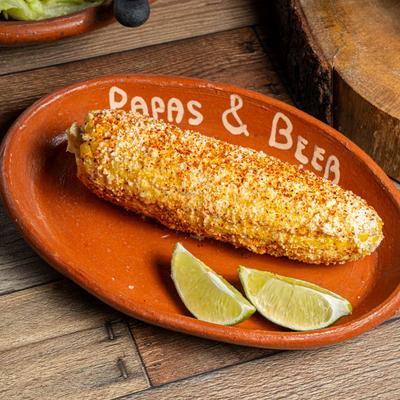 Mexican street corn on a clay plate labeled Papas & Beer, with lime wedges, on a wooden table.