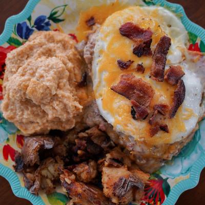Fried eggs over a biscuit topped with cheese and bacon, served with grits and crispy potatoes.