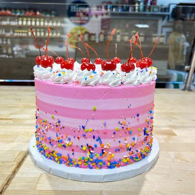 A pink cake with white whipped cream swirls topped with cherries and colorful sprinkles.