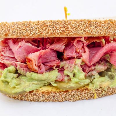 A sandwich with pastrami, creamy green guacamole, and pickles, served on seeded bread.