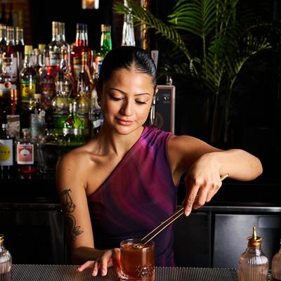 A bartender garnishes an amber cocktail served in a rocks glass on a copper bar top.