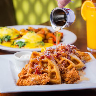 A person pours syrup on chicken and waffles, orange juice and Garden Benedict are in the background.