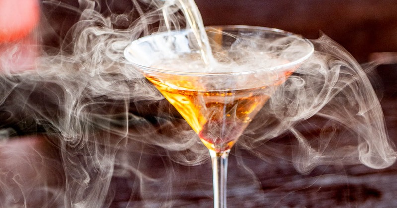 A martini glass with an amber liquid being poured in, surrounded by swirling smoke