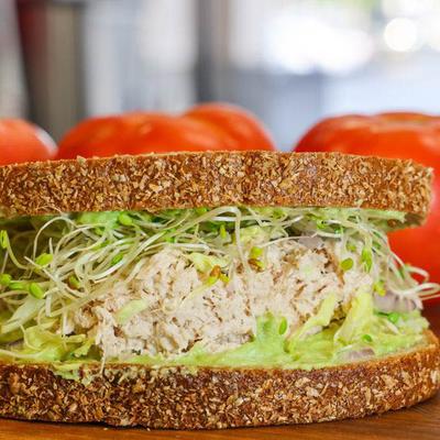 A sandwich with whole grain bread, tuna, sprouts, and avocado sits on a wooden board.