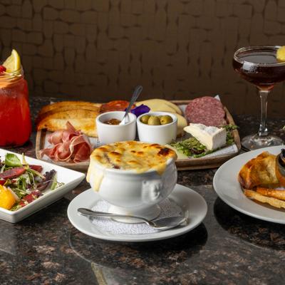 A French onion soup sits center stage among salad, charcuterie, and cocktails.