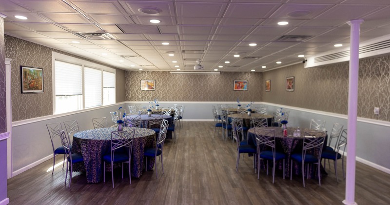 A banquet room with round tables and silver chairs, featuring wood floors and decorated walls