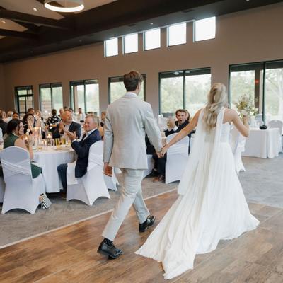 A wedding couple enters a reception hall, holding hands, Guests seated at  tables applaud warmly.