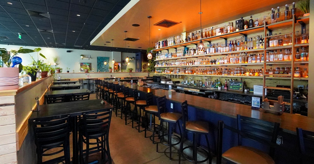 Interior, a long wooden bar top, black bar stools, shelves filled with liquor bottles,