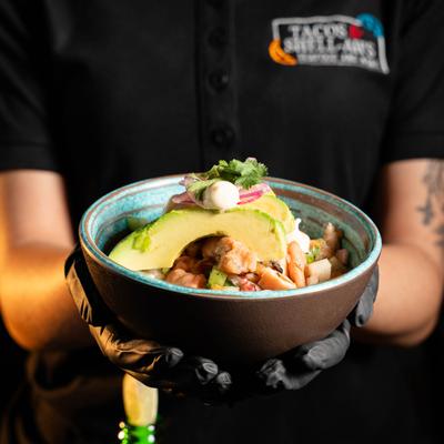 Gloved hands holding a bowl of fish ceviche topped with avocado slices.