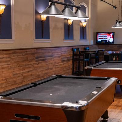 A dimly lit bar features two pool tables, wooden floors, wall sconces, and a small TV.