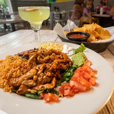 Seasoned chicken with rice, beans, tomatoes, and guacamole, accompanied by chips and a margarita.