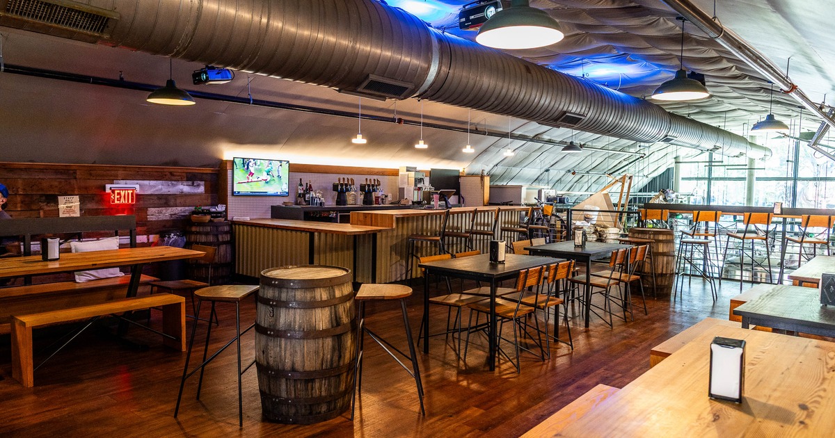 Rustic interior with exposed ductwork, wooden floors, tables, a central bar and a large window.