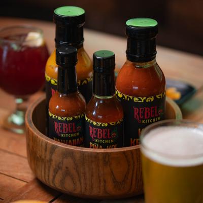 Hot sauce bottles in a wooden bowl beside a glass of beer.