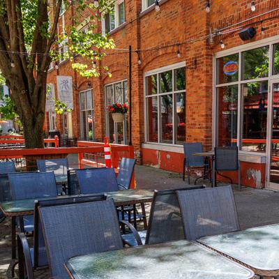 Outdoor dining area - tables, chairs, and a tree beside a red-brick building with large windows.