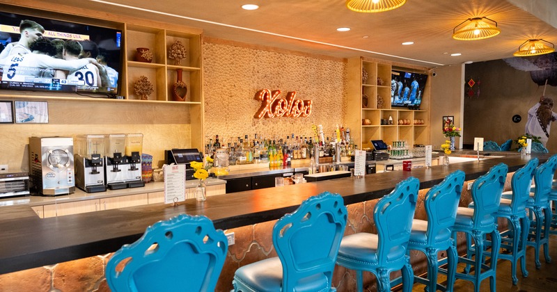 A large bar with blue chairs, a "Xolos" sign and a sports game on TV.