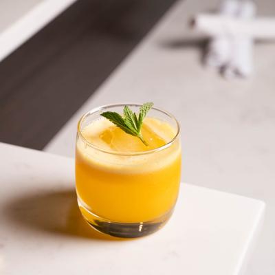 A glass of bright orange cocktail garnished with mint sits on a white marble surface,.