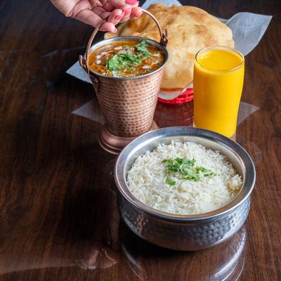 A copper pail with a lentil dish, a silver bowl of rice,  Mango Lassi, and naan on a table.