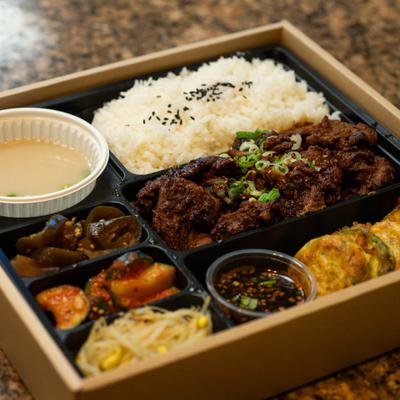 Korean lunch box featuring bulgogi beef, steamed rice, and various side dishes.
