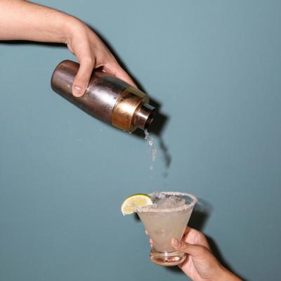 Cocktail being poured from a metal shaker into a salted-rim glass with ice and lime garnish.