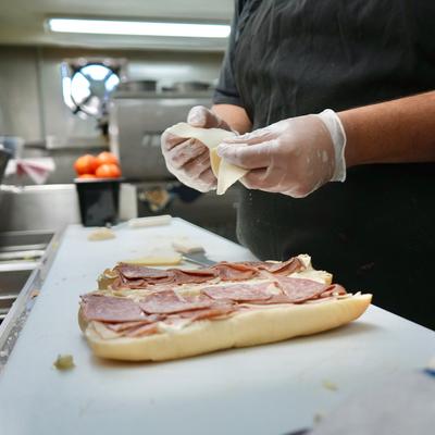 A person wearing gloves layering a cold-cut sandwich with deli meats and cheese slices.